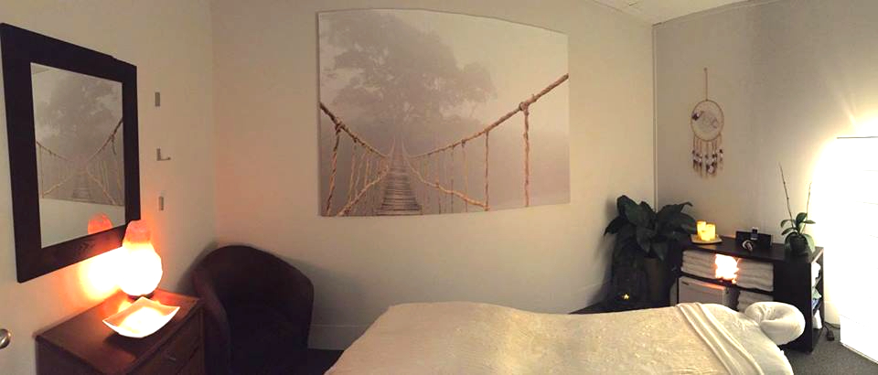 James's relaxing treatment room, with soft lighting, large photo print of rope bridge on back wall, a Buddha fountain sits on the floor in the far right corner. a soft padded massage table sits in the middle of the room on an angle covered in soft white linens and a cream fluffy blanket. a rounded back sitting chair is in the far left corner.